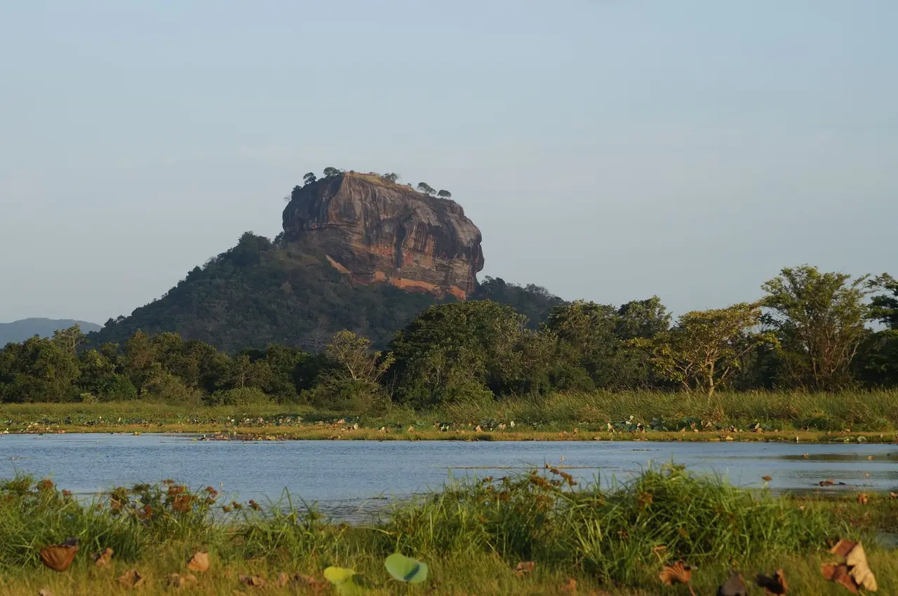 View of the iconic Sigiriya Rock Fortress surrounded by lush greenery and water, captured at sunrise.